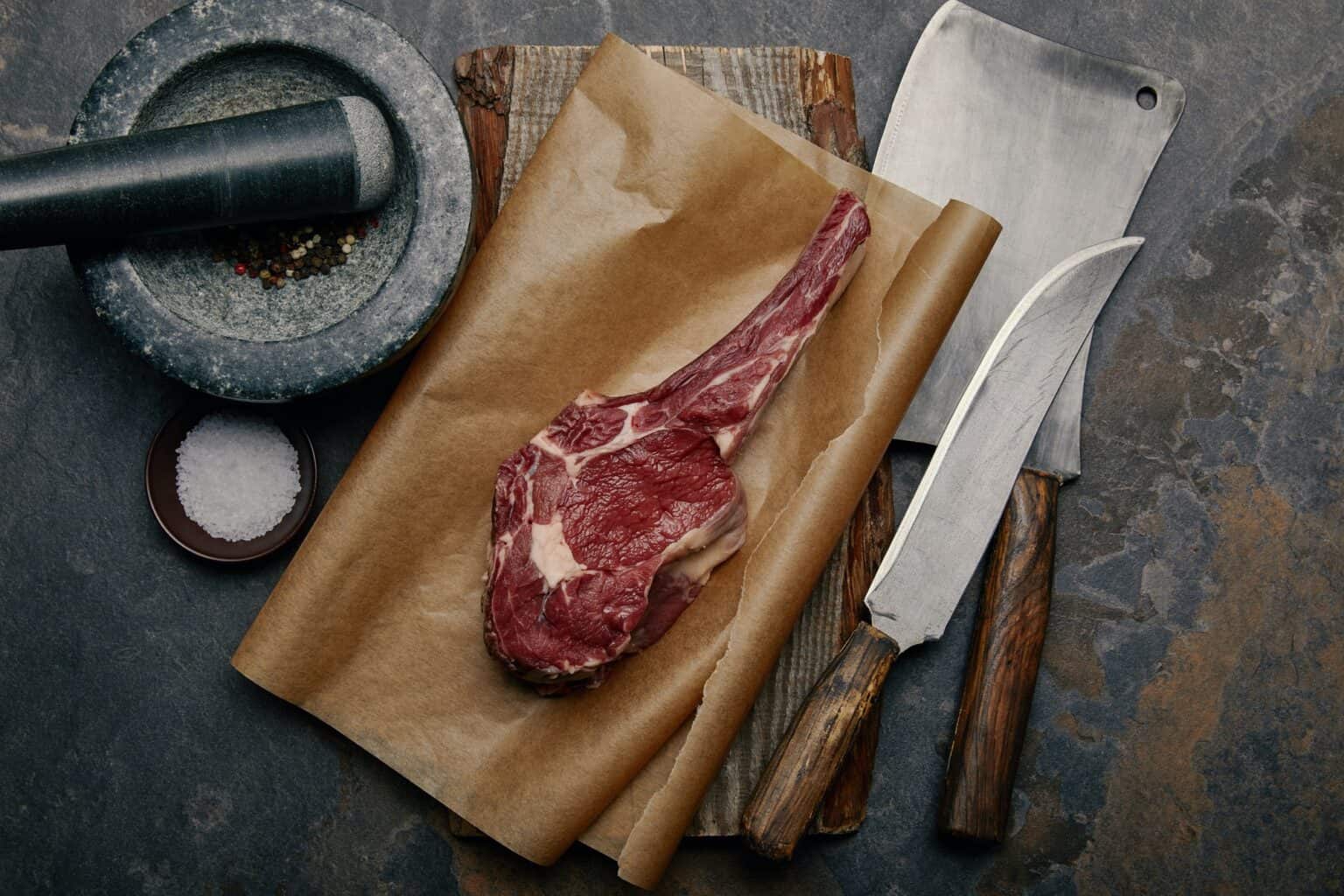 top view of raw rib eye steak on baking paper with kitchen knives and marble pounder on grey